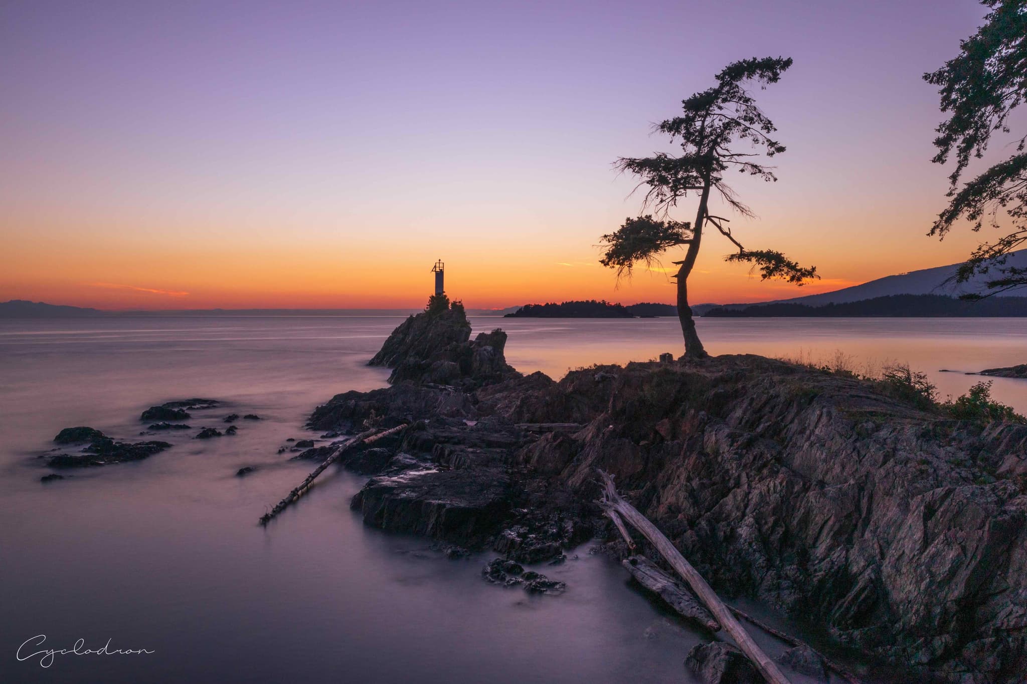 Coastal lighthouse on rocky outcrop at sunset with windswept tree and long exposure water